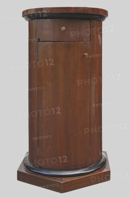 Cabinet-pedestal, message provenance, columns architectural motif, classicism and empire styles, early 19th century, possibly Northern Germany or Poland, piece of furniture storage, height 84 cm, diameter 46.5 cm., A cylindrical wooden cabinet in brown tones with a darker brown or black rounded trim near the top and base, sitting on a brown hexagonal wooden platform; the cabinet has a small round metal knob on a small top panel and another small round metal knob on a larger front door panel, with visible wood grain and a few small scuffs and lighter marks on the surface, set against a plain light gray background.