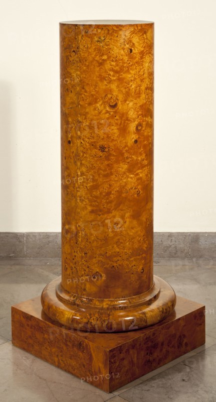 Pedestal, creator, provenance unknown. Biedermeier style. Pedestal in the shape of a column with a smooth shaft, a cylindrical base, on a square plinth. Entire piece veneered with birch burl. Shaft made of eight boards glued together at an angle from the front. Four plinth boards connected with diagonal dovetails. Shaft fastened to the plinth with four screws and additionally mounted on a central peg. Dated 1825–1835. Likely Poland. Furniture. Height 111.5 cm; width 44.5 cm; depth 44.5 cm., A tall cylindrical wooden column with a glossy, richly grained surface in warm amber and honey brown tones standing on a rounded wooden base and a square plinth of matching wood grain, set on a gray stone floor against a pale cream wall.