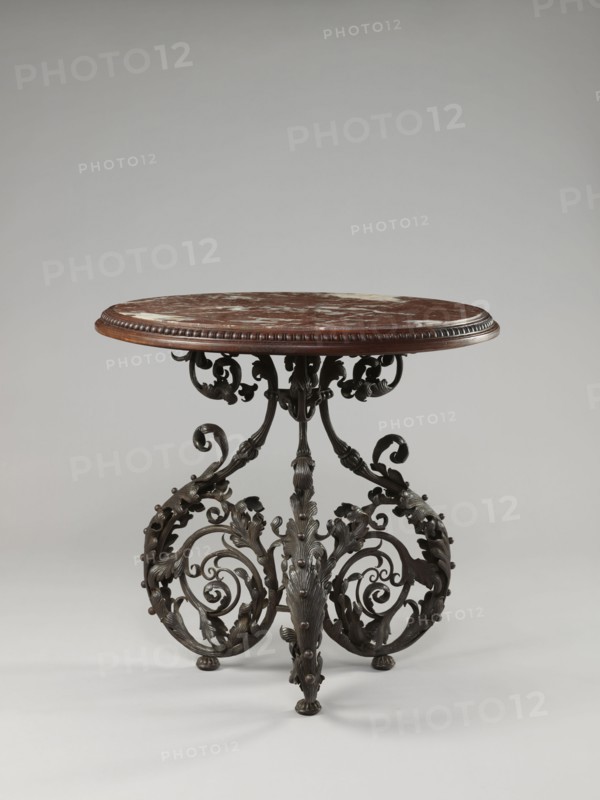 Table, Szewczykowski, Roman 1849–1901 author, plant decorations, plant twig, astragal, in the form of three S-shaped pieces, 1890s, Warsaw, furniture, table, height 80.7 cm, diameter 84 cm, A round table with a brown and white mottled top and a carved brown wooden rim stands on an ornate dark metal base; the base features a group of scrolling, leafy, and curling decorative elements with a dark bronze or black finish and small rounded feet, and the overall color palette includes brown, white, dark bronze or black, and hints of lighter gray from reflections.