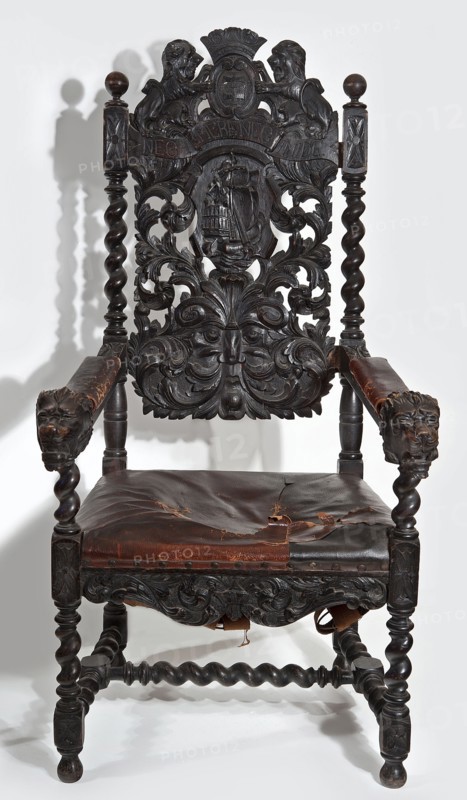 Armchair, origin unknown, neo-Baroque, early 20th century, Central Europe. Carved armchair with a padded trapezoidal seat and a high openwork backrest topped with the Gdansk coat of arms supported by two lions, with the motto NEC TEMERE NEC TIMIDE on the sash below. Straight, spirally turned legs with H binding joints. Straight, narrow frame, wider front, wavy-cut, carved with symmetrical acanthus twigs. Wide central backrest board with a cartouche rollwork with a sailing ship enclosed by a fleshy acanthus twig passing into a mascaron at the bottom; mounted between spiral side rails topped with spheres. Straight, angular armrests with padded rests culminating in a lion mask at the front. Structural elements join the cube. Frame painted black. Seat upholstery on springs, upholstered with brown leather, including the armrests. Resembling Gdansk Baroque. Furniture seating piece. Height 135 cm, width 69 cm, depth 75.2 cm., A dark wooden armchair with elaborate carved details fills the fram...