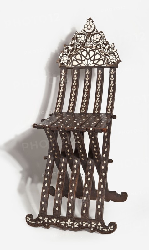 Chair with footrest, seat, and backrest composed of five slatted elements, richly inlaid with bone and mother-of-pearl with stylized plant patterns. Supports are connected to each other and to the seat with wooden pins enabling the structure to be folded and unfolded, terminating at the bottom with decoratively carved runners. The backrest is topped with a wide board with a wavy outline resembling a triangle. The slats are decorated on the front and sides with plant tendrils, topped with symmetrical geometric decoration at the bottom and a plant tendril with flowers at the top. The pins are topped with turned knobs. 2nd half 19th century, possibly Syria. Piece of furniture seating. Height 97 cm; width 44.2 cm; depth 57.2 cm. Materials: wood inlaid with bone and mother-of-pearl; geometric and plant decoration., A decorative wooden folding chair with dark brown wood and white inlaid floral and leaf patterns, featuring a carved ornate top with a fan-like lattice and flower motifs, a slatt...