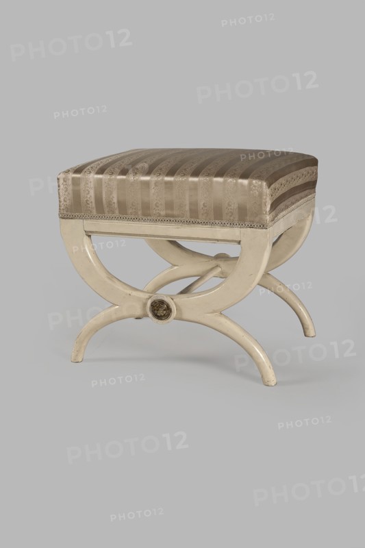 Stool, origin unknown, Empire style, rosette ornament. Rectangular stool with a black painted frame, upholstered, supported on the longer sides by crossed supports composed of two arched squares fastened in the middle and connected with a turned, simple link. At the intersections of the crosses are turned plaques with round fittings of embossed gilt brass sheet, each with a central rosette and a tendril on the edge. Padded seat with high sides, upholstered with non-original green striped fabric alternating grosgrain and satin. c. 1810, Central Europe. Furniture; seating piece. Dimensions: height 40 cm, width 43.2 cm, depth 35.3 cm., A small upholstered stool with a striped fabric seat in shades of beige and light brown, sitting on a curved wooden frame painted cream; the seat has a subtle patterned texture and a decorative trim along its lower edge, and the frame features intersecting curved legs forming an X-shape with a round decorative medallion at the center, all shown against a pl...