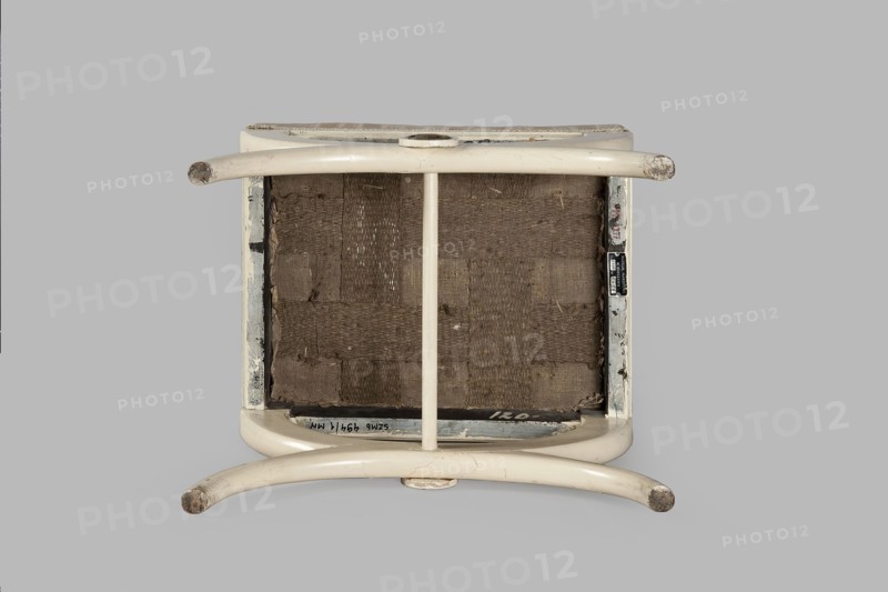 Stool, origin unknown, Empire style, rosette ornament. Rectangular stool with a black painted frame, upholstered, supported on the longer sides by crosses composed of two arched squares fastened in the middle and connected with a turned, simple link. At the intersection of the crosses are turned plaques with round fittings of embossed, gilded brass sheet, with a central rosette and a tendril on the edge. Padded seat with high sides, upholstered with non-original green striped fabric alternating grosgrain and satin. Circa 1810, Central Europe. Furniture, seating piece; height 40 cm, width 43.2 cm, depth 35.3 cm., An overturned stool with curved tubular legs and a central support, showing its underside with woven fabric webbing; the stool frame and legs are off-white or cream with areas of wear exposing darker spots, and the webbing is brown; the background is a flat light gray.