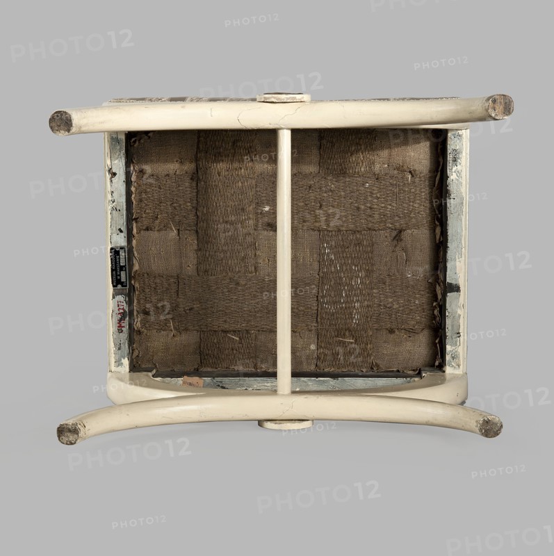 Stool, origin unknown, Empire style, rosette ornament. Rectangular stool with a black painted frame, upholstered, supported on the longer sides by crosses composed of two arched squares fastened in the middle and connected with a turned, simple link. At the intersection of the crosses are turned plaques with round fittings of embossed gilded brass sheet, with a central rosette and a tendril on the edge. Padded seat with high sides, upholstered with non-original green striped fabric alternating grosgrain and satin. c. 1810, Central Europe. Furniture, seating piece. Dimensions: height 40 cm, width 43.2 cm, depth 35.3 cm., An upside-down wooden stool or small chair with a cream or off-white painted frame showing scuffs and worn areas, a woven seat underside made of coarse brown fibers in a grid pattern, a central vertical support painted the same cream color, rounded legs with exposed worn ends, and a neutral light gray background.