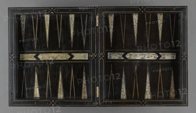 Chess, mill and backgammon box, games, flower vases, chess, plant decoration, baroque style, landscapes, backgammon, animals, birds, checkers party game, 1680s, France, furniture, equipment, game furniture, height 9 cm, width 37 cm, depth 32.8 cm, A rectangular hinged wooden board opened flat showing two panels with a symmetrical geometric pattern; the dominant color is dark brown to black in the field, with ivory-colored elongated triangular inlays and a few narrow pale lines creating pointed motifs; there are several horizontal pale rectangular inlays with darker decorative markings, and thin golden or yellow linear accents outlining panels and forming small diamond-like motifs near the center hinge; the outer frame is a darker wood with lighter edge lines and small circular holes along the border, and a metal hinge is visible at the center where the two panels meet.