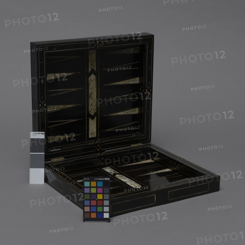 Chess, mill and backgammon box; maker, games; flower vases; chess; ; plant decoration; baroque style; landscapes; backgammon; animals; birds; checkers; 1680s, France; furniture; game furniture; height 9 cm; width 37 cm; depth 32.8 cm, A rectangular hinged box sits open on a gray background, revealing a shallow interior and a lid with geometric inlay. The box is predominantly black with decorative inlaid accents in a pale beige or ivory tone and thin gold lines outlining panels and triangular motifs. A few worn pale beige inlaid triangular shapes appear inside the lid and base. A small color calibration chart with multiple colored squares and a gray scale card stand near the front of the box, showing a range of colors including red, green, blue, yellow, purple, brown, black, white, and various grays. The overall scene includes shades of black, gray, pale beige or ivory, and gold, with small areas of brighter colors on the calibration chart.
