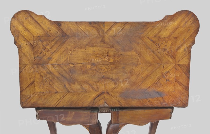 Game table, Rectangular with an extendable top square when extended with rounded, protruding corners, supported by four cabriole legs ending in hoof-shaped feet. Foot stained walnut; top veneered in walnut with inlay. Rear frame two-part: outer composed of two rotating frames on wooden hinges with legs attached on the inner side supporting the foldable top plate. Outer side of top framed by a crosswise frame with an asymmetrical, oval-shaped field, enclosed by channel bars and a ribbon, filled with floral and rocaille ornamentation. Inner side of tabletop has a wide walnut-veneered frame; center lined with green cloth. Game furniture, table; 1745–1770; Central Europe, possibly Saxony. Dimensions: height 74.6 cm, width 95 cm, depth 49 cm., A wooden tabletop seen from above with a warm brown and golden finish, showing wood grain patterns and a central decorative inlay with curved shapes and floral-like motifs; the tabletop has rounded corners on one side and straight edges on the other, ...