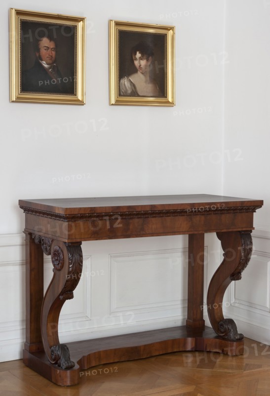 Console, Simmler, Jakub Jan 1791-1872 carpenter author, acanthus ornament, volutes, kimation ornament, rosette ornament, Biedermeier style. Rectangular console, supported by four supports the front S-shaped, widened at the top, the rear angular placed on a platform, veneered with mahogany, carved. The platform has a wavy cut at the front. The frame is straight, enclosed by a flap at the bottom, under the top processed with lesbian kimation with leaves with semicircular tips. The lower and upper volutes of the front legs are decorated with acanthus leaves on the front, and with rosettes on the sides. The rear edges of the front legs are enclosed by half-rolls. At the bottom of the side frames, between the legs, are decorative aprons made of acanthus twigs. The platform frame is braced with two additional slats. 1830–1840, Warsaw. Furniture; table. Height 88.4 cm, width 122.7 cm, depth 53.6 cm., A wooden console table with a rich brown finish and carved scroll legs stands against a white...