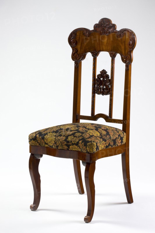 Chair, plant decoration, Louis Philippe style, acanthus ornament, mahogany chair with a padded seat and a high openwork backrest, with carved decoration. Square legs, S-shaped front legs with volute feet, rear legs arched back. Wavy frame with rounded front corners, seat in the form of an overlay cushion. Backrest framed on the sides by straight stiles extending the rear legs, topped with a board with a symmetrical, wavy-cut outline, emphasized by carved acanthus appliqués. The filling has two straight stiles covered with astragalus, topped and fastened together with acanthus leaf arrangements, and a narrow lower stile arched between the stiles. 1850–1860, Poland, possibly Warsaw. Seating piece furniture. Height 118.5 cm; width 51 cm; depth 52 cm., A wooden chair with a carved backrest and decorative central splat sits against a plain light background, the wood showing warm brown tones and a glossy finish; the seat is upholstered in a dark fabric with a floral pattern in shades of gold...