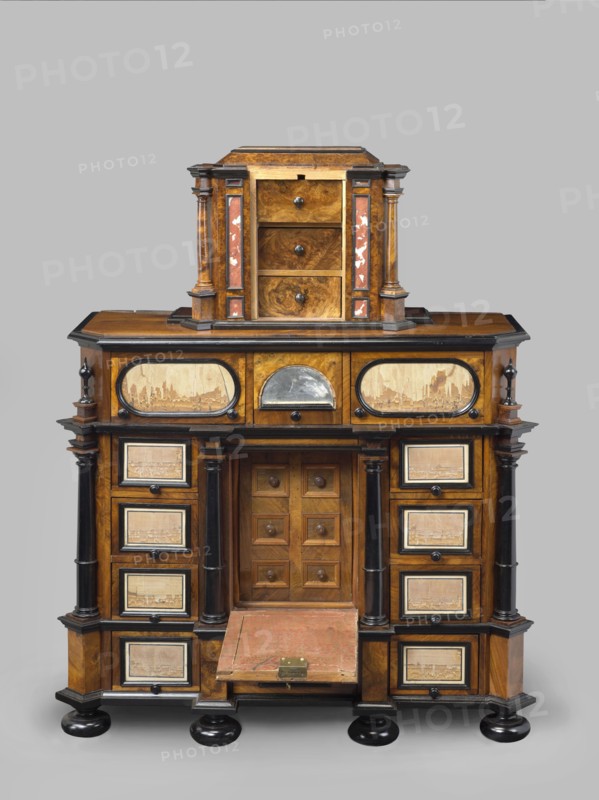 Cabinet, baroque, columns, birds, early 18th century, Southern Germany, possibly Augsburg, piece of furniture storage piece, height 114 cm, width 92 cm, depth 41 cm, creator unknown, provenance includes repossession., A wooden cabinet with a tiered form stands against a plain gray background, featuring warm brown wood tones with darker brown and black accents, and touches of reddish inlay on the upper section. The piece has a smaller upper chest with three drawers flanked by columns and decorative panels, sitting atop a larger lower case with a central recessed compartment behind a small door and a drop-front panel covered in worn red material with a metal latch. The lower section includes a group of drawers on each side framed by dark columns and rectangular panels that show lighter wood inlays with textured surface details. The cabinet rests on rounded black bun feet and shows polished wood grain, contrasting black trim, and small round wooden knobs on many of the drawers.