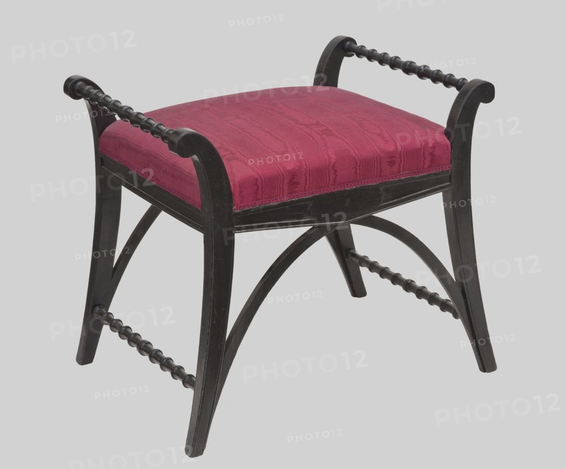 Gondola, Kolbinski, Jan Nepomucen 1820-post 1877, carpenter; neo-Empire style; GONDOLKA in the Empire style, on angular, arched legs flowing into openwork stiles of channel-shaped sides turned outward and ending in a volte, with a padded rectangular seat. The frame is stained black and polished. The side leg joints and armrests are spool-turned; the front and rear joints are arched slats forming a pointed arch. The frame is flanked at the bottom, widening in the long middle section, in the outline of a flat triangle pointing downwards. Seat upholstery on straps, padded with grass, upholstered with silk-cotton jacquard fabric with palmettes arranged in a net pattern red plain weave background; patterns created with protruding threads with metal braiding, finished with narrow galloon with metal braiding thread. 1877, Warsaw. Furniture; seating furniture. Height 49.4 cm; width 57 cm; depth 43 cm., A small upholstered stool with a rectangular cushioned seat covered in deep red fabric showi...