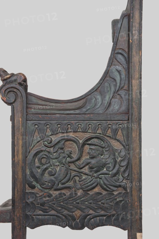 Armchair, wickerwork ornament, battle scenes, knights, plant ornament, dogs, ca. 1500 or 1st half of the 16th century, Sweden, Norway, piece of furniture, seating piece, height 112 cm, width 75 cm, depth 58 cm, The image shows a carved wooden panel with intricate raised reliefs and decorative molding, featuring a central human-like figure with flowing hair and a headband, seated or reclining and interacting with a stylized animal or serpent-like creature through a curved, ribbon-like form; surrounding the scene are swirling leaf and wave motifs, braided patterns, and scalloped edges. The wood displays a worn, aged surface with visible chips and scratches, and the predominant colors visible are dark brown and deep green with hints of muted reddish-brown where the finish is worn.