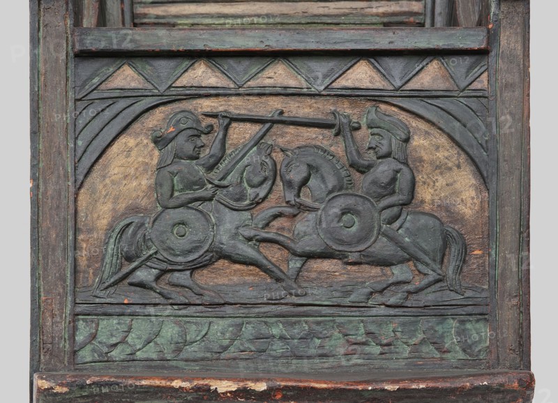 Armchair, wickerwork ornament, battle scenes, knights, plant ornament, dogs, ca. 1500 or 1st half of the 16th century, Sweden, Norway, piece of furniture, seating piece, height 112 cm, width 75 cm, depth 58 cm, A carved wooden relief panel shows a symmetrical scene of two mounted figures facing each other on horseback, each holding a long spear raised toward the other and carrying round shields at their sides; the riders wear distinctive helmets and have visible facial features and limbs, and the horses are rendered with bridles, manes, and raised legs suggesting motion. The carving is set within a rectangular frame with a curved arch above the scene and geometric triangular motifs in a band above; a carved leafy pattern runs along the bottom border. The surface shows layered paint and worn patina with visible colors including green, dark gray, brown, muted gold, and small areas of reddish-orange where the underlying material shows through.