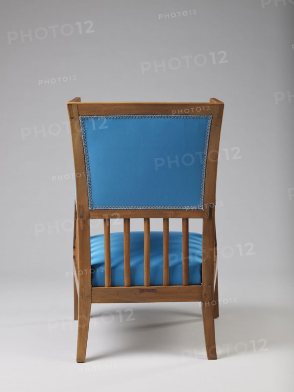 Armchair Berzera, pre-war collection. ARMCHAIR-BERZERA with a slender backrest and sides equal in height, arching upwards and forming ears, with angular frame elements. Front legs converge, rear legs arch outwards. Narrow, straight frame; trapezoidal seat in the form of a removable cushion on springs. The upper part of the sides and the backrest, which is slightly wider in the upper part, are upholstered, the lower part is filled with vertical slats. The frame elements are additionally connected with single pegs. Ca. 1820. Possibly Warsaw, Vienna, or Lesser Poland. Piece of furniture, seating piece. Height 86.5 cm, width 54.8 cm, depth 61.8 cm., A wooden chair viewed from the back with a rectangular upholstered back panel in bright blue trimmed with decorative edging, a matching bright blue cushioned seat visible through vertical wooden slats, and a light brown wooden frame and legs set against a neutral light gray background.