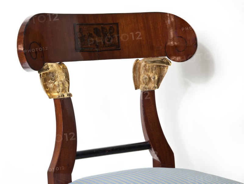 Chair, furnishings of representative buildings of the Republic of Poland 1918–1939, Administration of the Imperial Palaces in Warsaw 1815–1915, eagles, plant decoration, landscapes, Empire style. Mahogany chair with a padded seat removable cushion and an openwork backrest topped with an arched pelta-shaped board supported by a pair of eagles. The front legs are club-shaped, with the upper section carved with acanthus leaves; the rear legs are angular, curved outward in an arched manner. The seat and frame are straight at the front, arched at the back. The backrest rails are an extension of the rear legs, one-third of the height is arched inward, fastened at the bottom with a turned black link. Early 19th century, Warsaw; seating furniture; height 83.1 cm, width 48 cm, depth 52 cm., The image shows the upper portion of a wooden chair with a curved top rail that has rounded scroll ends and a rectangular inset panel; the chair back has two carved vertical supports topped with gilded decor...