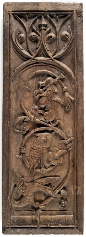 Furniture panel in bas-relief in the shape of an elongated standing rectangle, late 15th century, France. Fragment of furniture height 72.8 cm, width 25.2 cm, depth 3 cm. Decoration: plant twig with flowers bearing a dragon, an owl, and an animal playing bagpipes possibly a dog or monkey sitting on it; at the top tracery with fish-bladder motifs and a lily flower. Decoration enclosed in a rectangular frame with lines intersecting in the corners. Gothic style; drôlerie; animals, fantasy animals, plant ornament, musical instruments., A tall rectangular wooden panel carved with a vertical sequence of decorative motifs, showing a range of carved forms in brown wood tones. At the top a rounded arch framing stylized leaf and vine shapes with curved, pointed elements. Below that an arched frame contains a carved animal figure sitting upright holding an implement, with surrounding curved branches and floral motifs. Further down another circular frame encloses a carved bird with detailed feathe...