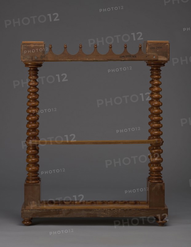 Pipe stand, origin unknown, smoking, pipes, acorns, rack for pipes and billiard cubes. Skeletal structure composed of two pairs of spool-turned columns on angular plinths, mounted on a narrow rectangular platform supporting cuboid drawer bodies, connected mid-height and at the top by horizontally arranged slats with semicircular cutouts on the front. Platform rests on four turned legs with a high straight rim, with glued-on wavy-cut slats for holding billiard cues or long pipes, and a single groove at the back. Upper panel decorated with a gallery in the form of an arched crenellation reverse arcading topped with acorns. Bodies with drawers probably intended for balls are surrounded by a low slatted gallery. 1830–1840, Central Europe. Furniture/equipment. Height 111.5 cm; width 89 cm; depth 29 cm., A wooden freestanding frame with a dark brown finish standing against a neutral gray background, featuring two turned vertical posts with a repeating bulbous profile connected by a horizonta...