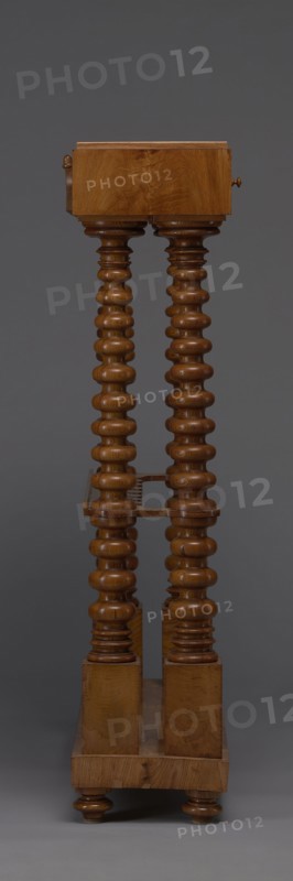 Pipe stand, origin unknown, smoking accessory featuring pipes and acorns; rack for pipes and billiard cubes. Skeletal structure of two pairs of spool-turned columns on angular plinths mounted on a narrow rectangular platform, supporting cuboid drawer bodies connected mid-height and at the top by horizontal slats with semicircular cutouts on the front. Platform rests on four turned legs with a high straight rim and glued-on wavy-cut slats for holding billiard cues or long pipes, with a single groove at the back. Upper panel decorated with a gallery in the form of an arched crenellation reverse arcading topped with acorns. Drawer bodies probably intended for balls are surrounded by a low slatted gallery. Dated 1830–1840, Central Europe. Furniture/equipment. Dimensions: height 111.5 cm, width 89 cm, depth 29 cm., A tall wooden piece of furniture with a warm brown finish stands against a dark gray background; it has a rectangular top block and a rectangular base connected by a pair of orna...