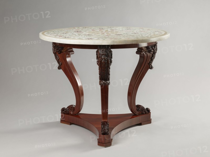 Table by Jakub Jan Simmler 1791–1872, carpenter. Round table supported by three S-shaped carved legs with acanthus volutes at both ends, mounted on a triangular platform with concave sides. Legs strongly flared at the top with half-cylinders at the rear edges, topped with volutes. Platform has three flat, block-shaped feet. Round, narrow frame glued from four layers of slats into a brick shape. Ornament: acanthus, plant decoration, volutes. Style: Louis Philippe. Function: furniture, table. Date: 1840–1847. Dimensions: height 76.4 cm; diameter 93.5 cm., A round table with a light-colored patterned top rests on a dark wooden base; the tabletop shows muted tones including pale, greenish, and reddish hues in a decorative design, and the edge appears creamy. The wooden base is a rich brown with a polished finish and features three curved legs joined to a triangular platform, each leg carved with ornate floral and scroll motifs in darker brown tones. The background is a smooth, neutral gray...