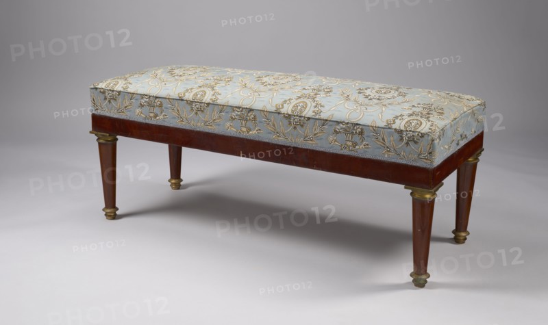Banquette, Empire style, early 19th century, Warsaw, rectangular mahogany banquette, upholstered, supported by four turned, tapering legs with brass capitals and feet. Straight polished frame with cast, chiseled, gilded fittings. Spring-mounted upholstery. Dimensions: height 52 cm, width 122.5 cm, depth 48 cm., A rectangular padded bench with a patterned fabric top in pale blue and gold rests on a wooden frame in warm brown; the fabric shows ornate floral and medallion motifs in gold on the blue background. The wooden skirt is brown and the tapered legs are brown with gold-colored metal caps and collars. The bench sits on a smooth light gray surface against a neutral gray backdrop, and there are soft shadows beneath the bench. The visible colors include pale blue, gold, brown, light gray, and neutral gray.