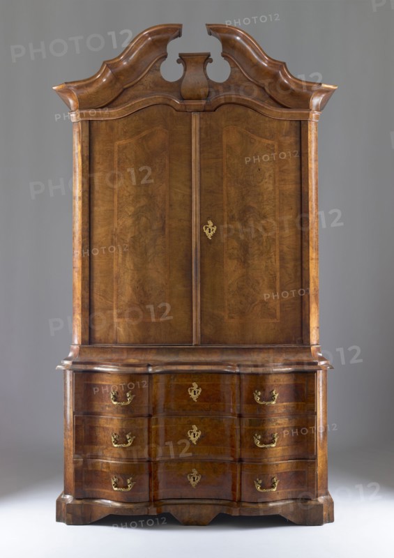 Sideboard, gift, architectural elements, plant decoration, two-part sideboard with chamfered and concave front corners, wavy front and straight sides, three-drawer base and top closed with double doors, supported on console legs; veneered with patterned walnut, inlaid with ribbon motifs, polished. The tops of the upper section are framed by richly profiled cornices, the upper one with a flat roof and a volute cornice with a central corbel. The top leaves have a wavy design creating a crossbow-like outline, as does the narrow cornice above them. The sides of both bodies, and the doors and drawer faces, are framed with a ribbon and cross-shaped frame. Three shelves not original are inside. Construction of both bodies, drawer faces, and door leaves is board-like; the back is framed with two panels; bases are board-like. Rims fastened to sides with wedges. Steel applied locks; single-bolt drawer locks; double-bolt door locks. Steel pintle hinges; single-bolt steel-brass latches. Brass pull...