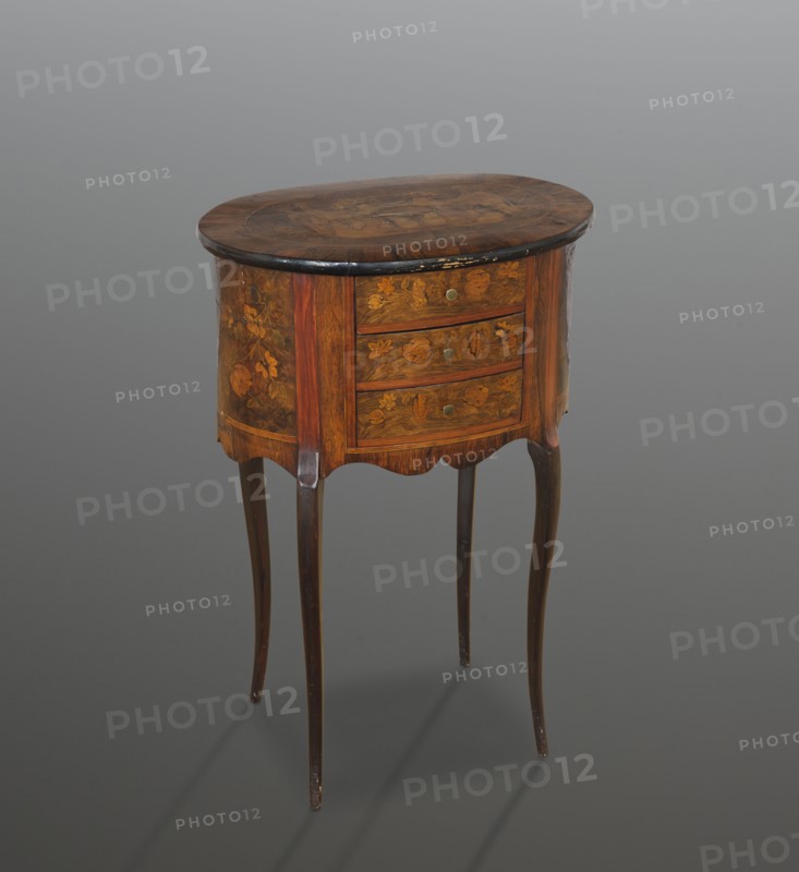 Chest of drawers table, classicism and rococo styles, floral decorations. Oval chest with a tall body containing three drawers, supported by four slender S-shaped legs, richly inlaid. Body is wavy-cut at the bottom. Wooden top with a wide rosewood frame and quadruple veneer; oval field filled with a heraldic composition central coat of arms with a palm tree supported by standing lions, against a cloak suspended from two open five-stick crowns and flanked by a pair of Indians in plumes. Edge of the tabletop is rounded and blackened. Drawer handles are brass knobs. 4th quarter of the 19th century, Central Europe. Piece of furniture storage. Height 75.24 cm; width 50.1 cm; depth 34.4 cm., A small wooden oval-topped table with a dark brown top and lighter brown body stands on four slender curved legs in a darker brown. The front and sides of the body show decorative floral inlays in shades of brown and golden brown. The table has a scalloped apron beneath the body and a few round metal-col...