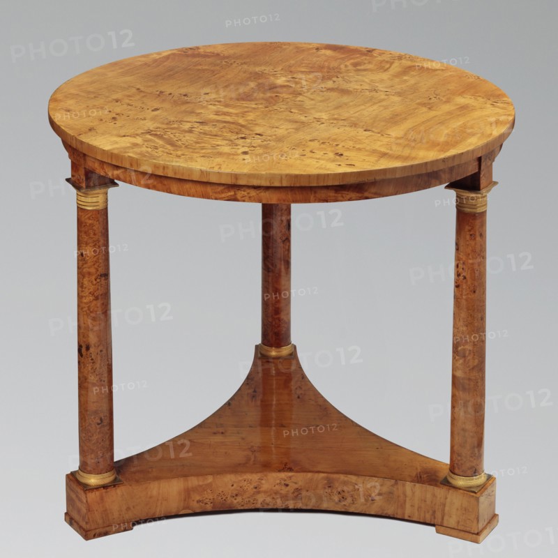 Round table on three legs in the form of slender columns with smooth shafts, mounted on a triangular platform with concave sides. Entire piece veneered with poplar burl. Cube feet at the corners of the platform. Bases and capitals of the columns are cast brass, covered with a twisted rope motif and stylized leaves. Biedermeier style, 1815–1825, Warsaw. Piece of furniture; height 75.8 cm, diameter 84 cm., A round wooden table with a warm golden-brown surface and visible wood grain sits on three cylindrical legs that have a slightly darker brown wood tone and natural wood patterning; a triangular wooden stretcher in a medium brown connects the legs and rests on a curved wooden base piece that matches the tabletop's golden-brown color, and there are small decorative bands of pale gold at the top and bottom of each leg.