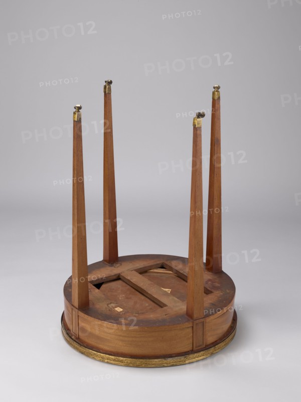 Table, classicism style, geometric ornament, round mahogany table with a white marble top enclosed by a brass gallery, supported by four slender, straight legs tapering downwards. Entire table is polished. Legs have knuckles at the top and brass rollers with angular clamps at the bottom. Drum-shaped frame contains a drawer with a lock. Stone slab is chiseled on the underside. Upper frame supporting the top is chiseled at points of varying thickness. Cast gallery gilded with amalgam. 1790–1810, France . Furniture, table. Height 77 cm; diameter 57.6 cm., A wooden round table shown upside down with a circular base in brown wood and a decorative gold-colored trim along the bottom edge, a few tapered wooden legs attached upright to the base with small gold-colored metal fittings at their tops, the wood showing natural grain and some darker worn areas, and a plain light gray background.