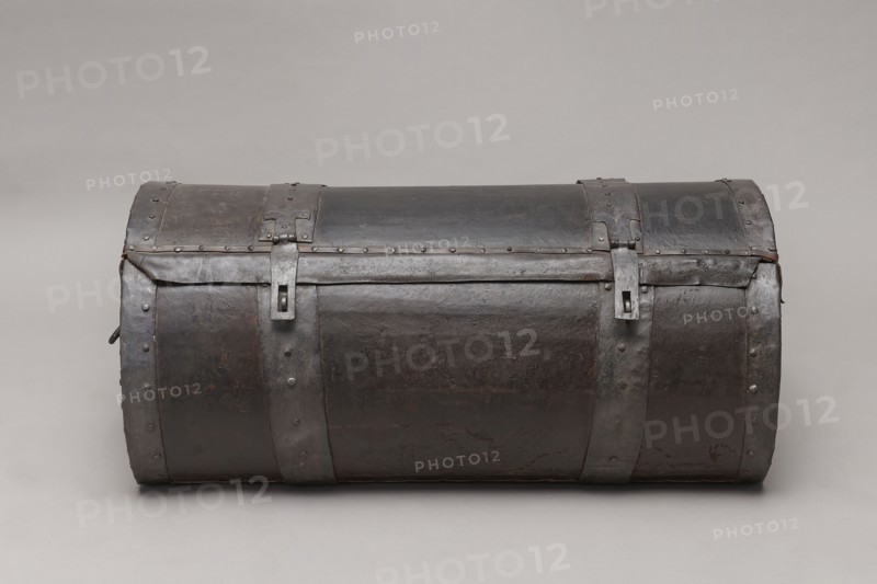 Trunk, A TUBE in the shape of a cylinder truncated at the base, covered with thick leather and upholstered with four steel straps, fastened with steel heads, with U-shaped handles on the sides of the body. The lid is closed with two hasps, attached to strap hinges. The connection between the body and the lid is protected from water by a leather apron. The side straps on the outside have semicircular cutouts, folded. The interior of the trunk is lined with gray canvas. The lid, when opened, is held on the sides with leather strips. 17th/18th century, Poland, piece of furniture, height 46 cm, width 104.5 cm, depth 51.5 cm., A cylindrical chest with a domed lid made of dark metal with visible rivets and metal bands, showing a worn and slightly textured surface in shades of dark brown and black with areas of dull gray; a few vertical metal straps wrap around the body and lid, each secured with rivets and featuring rectangular metal fastenings, and the piece rests on a smooth light gray bac...