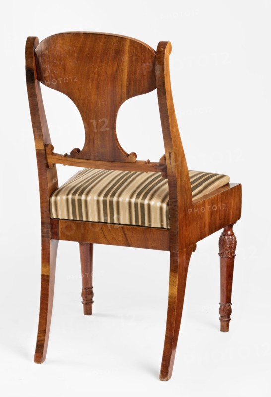 Chair, acanthus ornament, Biedermeier style, palmettes ornament. Front legs straight, turned in the form of a baluster; rear legs sabre-shaped. Trapezoidal seat, slightly bulging at the front. Wide side frames frame the seat cushion. The cushion rests on a pine frame, upholstered in green-striped fabric. Backrest composed of angular S-shaped side stiles, a lower crossbar, and a rounded central board placed between them, narrowed at the bottom, straight in line with the back. Backrest stiles widened in the lower section, ending at the top with a forward-curved volute. Polished frame enriched with woodcarving — acanthus leaves on the upper part of the front legs, antemion and symmetrical S-shaped beams at the bottom of the backrest boards, scrollwork at the ends of the backrest crossbars. 1830–1840, Warsaw, Poland. Seating furniture, furniture; height 92.3 cm, width 51 cm, depth 56.5 cm., A wooden chair shown from the back and side with a warm brown finish, a curved backrest with cutout ...