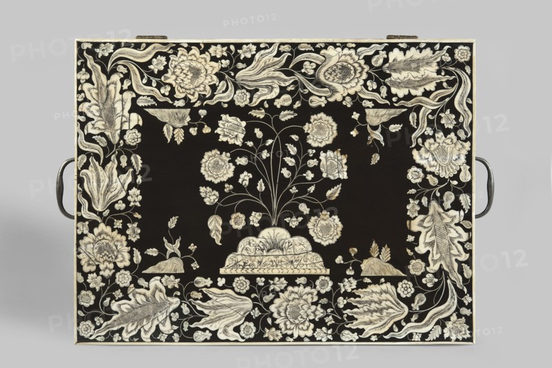 Travel dressing table, flowers, plant ornament, Baroque style, Lubomirski family, flower bushes ornament. Travel dressing table in ebony, cuboid, with a lifting lid and a drawer at the bottom of the front, richly inlaid with flowers and plant twigs in engraved bone, set with black paint. Inside: a tilting mirror, five compartments on each side six with lids. The mirror frame and lids are inlaid with bone with plant and floral twigs. Three compartments in the drawer. Steel handles on the sides swan neck. Ca. 1750, Vizagapatam, India. Travel/hygiene furniture. Height 17 cm, width 41 cm, depth 30.5 cm., A rectangular object with a black surface decorated with an intricate cream-colored floral pattern fills the image; a central stylized plant with stems and blossoms rises from a scalloped base, surrounded by a border of large ornate flowers, leaves, and curling tendrils, with a pair of dark metal handles visible at the left and right edges.