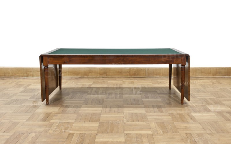 Table, Biedermeier style; rectangular table with drop-down flaps on the short sides; supported by four turned, tapering legs with knuckles at the top. Main top plate lined in the middle with grey cloth, framed by a mahogany frame; connected to the frame by sideboards. Flaps veneered with mahogany. Wooden sliders with slatted flap supports glued to the underside of the main top part on the short sides. Profiled legs with a roll-up at the top and pairs of rings midway; frame stained mahogany, polished. Inscription/association: Pilsudski, Józef 1867–1935. Date: 1830–1837. Place of origin: Warsaw. Function: furniture/table. Dimensions: height 78.4 cm; width 221 cm; depth 96 cm. Creator: unknown., A wooden table with a green top sits on a patterned wooden floor against a pale wooden baseboard and white wall; the table has a rich brown finish with turned legs and drop leaves on either side, and the visible colors include brown, green, tan, beige, and white.