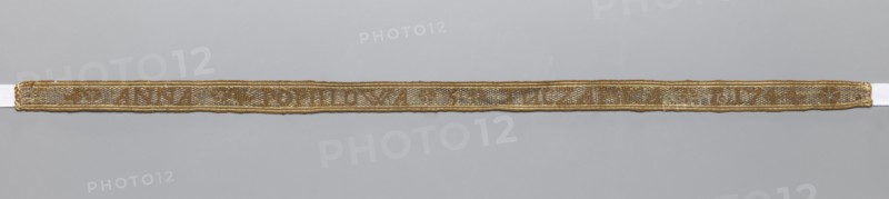 Gallon, galloons haberdashery; woven inscription: brown letters on a silver background; parallel to the edge, two white-brown stripes on each side; 1744; Poland; fabric; haberdashery; width 1.4 cm; length 41 cm., A narrow woven band lies horizontally across a pale gray background, showing a textured surface with a metallic sheen; the band has a warm golden color with areas of muted brown and pale beige, and it is edged with a slightly darker golden border.