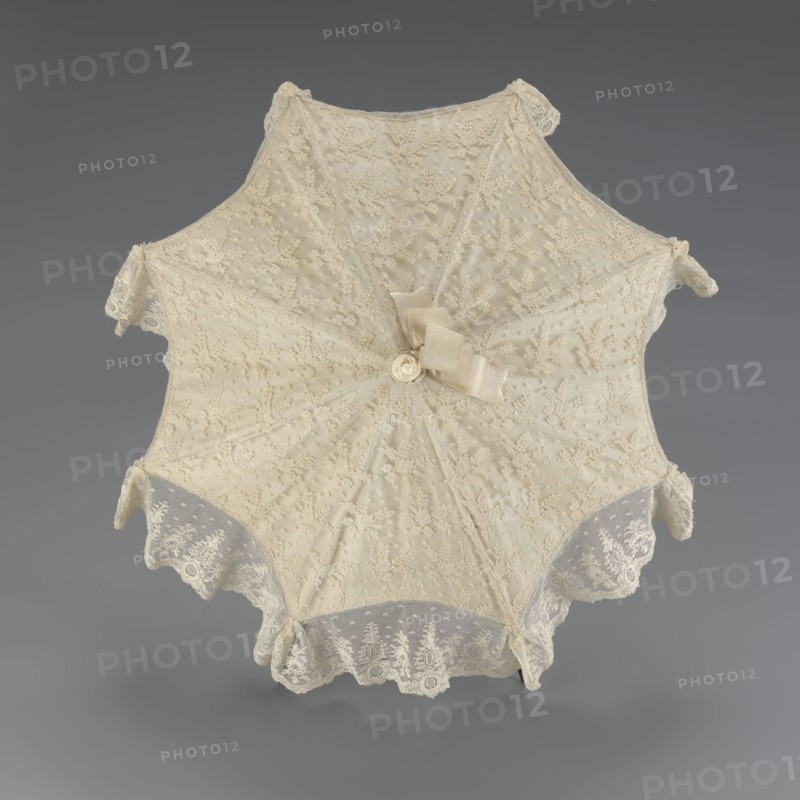 Umbrella, women's umbrella made of white lace. Canopy trimmed with cream silk at the bottom, concealing the metal structure and providing a backdrop for the lace when unfolded. Bone handle carved with floral motifs and folds in half. c. 1850–1875, France. Length 66 cm; diameter 57 cm. Categories/subjects: umbrellas, fashion, lace, women's fashion, 19th-century fashion accessories., A delicate lace parasol is shown from above, with an octagonal shape and scalloped edges, made of fine off-white and cream lace with floral embroidery and dotted mesh; a small bow and fabric rosette in similar cream tones sit near the center, and several lace ruffles and decorative tabs extend from the edges, all photographed against a smooth neutral gray background.