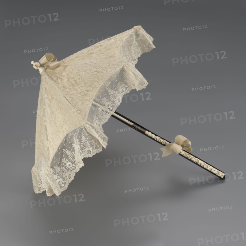 Women's umbrella made of white lace. Its canopy is trimmed with cream silk at the bottom, which conceals the metal structure and, when unfolded, provides a backdrop for the lace covering. The bone handle is carved with floral motifs and folds in half. c. 1850–1875, France. Length 66 cm; diameter 57 cm. Materials/attributes: umbrellas, fashion, lace, women's fashion, 19th century fashion accessories., A delicate lace parasol in shades of cream and ivory rests against a neutral gray background, featuring a scalloped lace canopy with floral and dotted patterns, a few decorative bows in a pale beige attached near the top and along the handle, and a slender dark shaft with a carved or inlaid lighter decorative band running along part of its length; the lace edges form a ruffled trim and the overall appearance is ornate and finely detailed.
