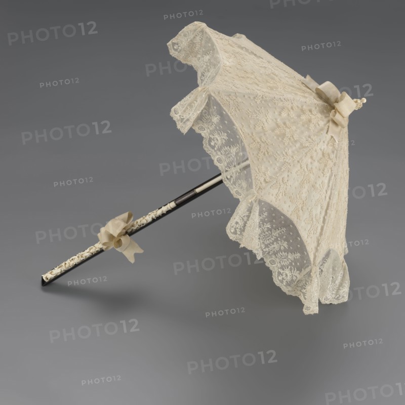 Women's umbrella made of white lace. Its canopy is trimmed with cream silk at the bottom, which conceals the metal structure and, when unfolded, provides a backdrop for the lace covering. The bone handle is carved with floral motifs and folds in half. c. 1850–1875, France. Length 66 cm, diameter 57 cm. Fashion accessory; 19th-century women's fashion., A lace parasol with an open canopy rests on a smooth gray background; the canopy is an off-white or cream color with delicate floral lace and scalloped edges, and it has a matching cream bow near the top and a few cream ribbons tied along the shaft; the shaft is dark, appearing black or very dark brown, with an inner section that shows a lighter, carved or textured cream-colored detail; overall colors visible include cream, off-white, black or dark brown, and gray.
