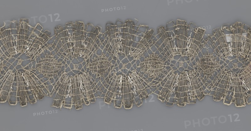 Lace fragment, lace, provenance unknown, metal threads, 18th century, Europe, lace fabric, width 8.2 cm, length 27.5 cm., A close-up of a decorative lace trim displayed against a flat background, showing a repeating scalloped pattern with open net-like sections and fan-shaped motifs; the lace appears metallic with a pale gold or silver sheen and some off-white threads, while the background is a muted gray.