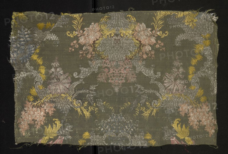 Fragment of fabric, fabrics, fragments of silk patterned fabric with floral patterns. Pattern with a symmetrical composition of fantasy-floral motifs: lilac branches entwined with silver leaves stylized into a Rococo ornament. Background: gros de Tours green, pattern brocaded with threads tied in a weft weave: lemon, celadon, light and dark pink, white, silver braided thread, brown. Piece width: 54.5 cm. Border width: 0.6 cm. 1st half 18th century, France; height 54.5 cm, width 36 cm., A rectangular fabric fragment with a muted olive-green background displays an ornate symmetrical floral and foliate pattern in a few colors including pale pink, soft gray, muted yellow, and a faded blue. The woven surface shows a central elaborate cartouche-like motif flanked by scrolling acanthus shapes and stylized blossoms, with clusters of rounded flowers and leafy sprays arranged across the cloth. The edges are slightly frayed and the weave texture is visible, with areas of metallic sheen in the gra...