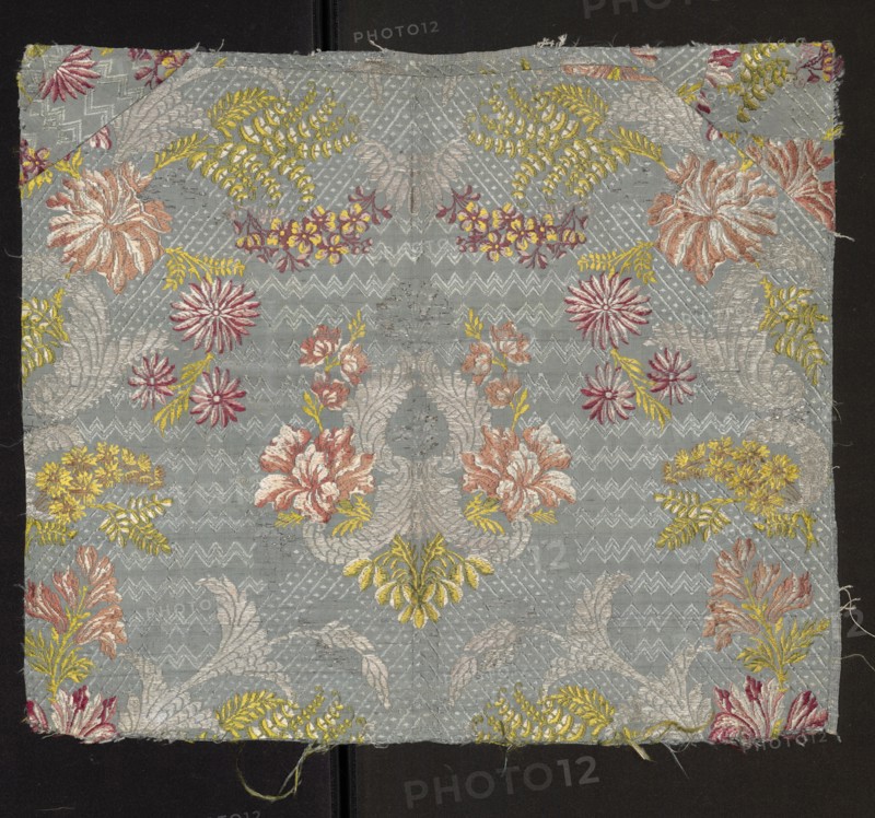 Fabric fragment: silk patterned with a symmetrical composition of branches of various stylized flowers surrounded by a geometric-floral overprint ornament. Blue background Gros de Tours, liseré; pattern brocaded with colorful threads in light green, yellow, white, blue, light and dark red, light brown, pink. Size of the repeat larger than the preserved fragment. Dimensions: length 38 cm report larger than fragment, preserved panel height 51.5 cm, width 44 cm; edge width 0.6 cm. 18th century, France., A square piece of woven fabric with a pale bluish-gray background covered in an ornate floral and foliate pattern in colors including pale pink, dusty rose, deep pink, yellow, gold, and silvery-white, with a repeating arrangement of stylized flowers, fernlike leaves, and curved acanthus shapes; the fabric shows surrounding raw edges and slight fraying, and it is placed against a dark background with a narrow vertical seam visible beneath the fabric.