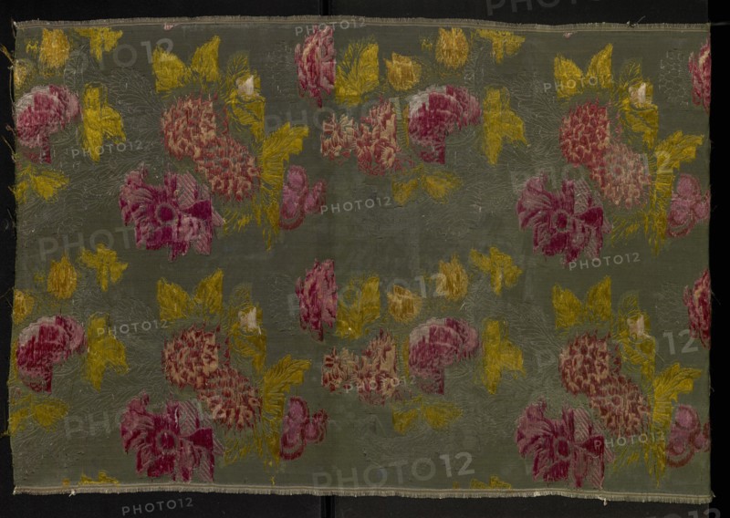 Fragment of patterned silk fabric, plant motifs in a naturalistic style, brocaded with tied weft threads in light and dark green, yellow, light and dark salmon and raspberry pinks, white, silver braided thread, and dark brown/black. Background: cannelé 3 coups with an effet poil traînant. Weave structure: Gros de Tours with effet poil traînant, liseré, broché à liége repris type 3. Rich use of silver threads enhances a strong chiaroscuro modelling reminiscent of Jean Revel's flower-and-fruit designs; three-dimensional brocade effect typical of Lyon silk weaving from the 1730s. Stiff silk with grosgrain structure, used for representative court dress Robe à la française in the first half of the 18th century. 18th century, France. Height 97 cm, width 55 cm., A rectangular piece of fabric with a muted green background displays a repeating floral pattern in shades of pink and deep rose for the blooms, mustard yellow for leaves and accents, and pale cream highlights; the surface shows woven ...