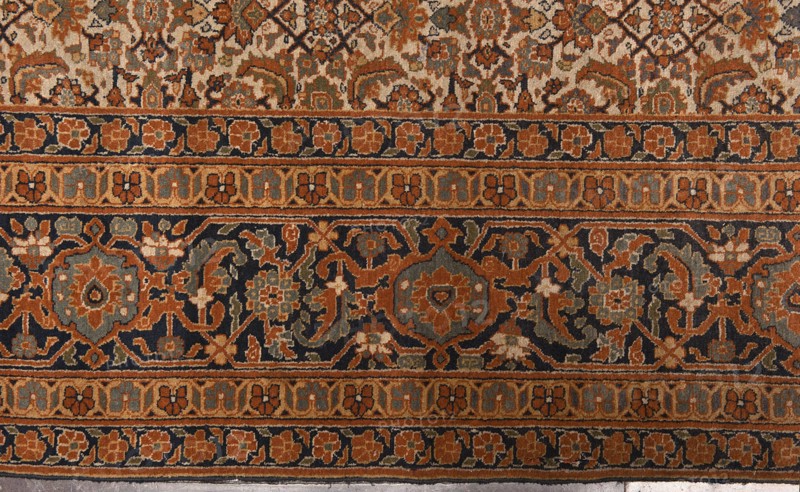 Carpet, 19th century, Iran. Knotted wool carpet with decorative herat-style pattern. Central field on a light cream background with a fine net ornament herat sack composed of lanceolate leaves, rosettes, palmettes, stylized flowers, and rhombuses. Five-band border with floral motifs; widest central band on a navy blue background. Colors: brick red, red, green, blue, navy blue, and white. Function: court and home furnishing. Size: height 520 cm, width 395 cm., The image shows a patterned textile with ornate repeating designs and horizontal bands. The dominant colors visible are rust orange, navy blue, beige, olive green, and muted gray-blue. There is a central dark band filled with floral and geometric motifs in orange, gray-blue, olive, and beige set against a navy background, flanked by narrower bands of small stylized flower shapes in orange and beige on navy and beige backgrounds. Above and below these bands are more intricate areas of interlocking floral and vine patterns in beige,...