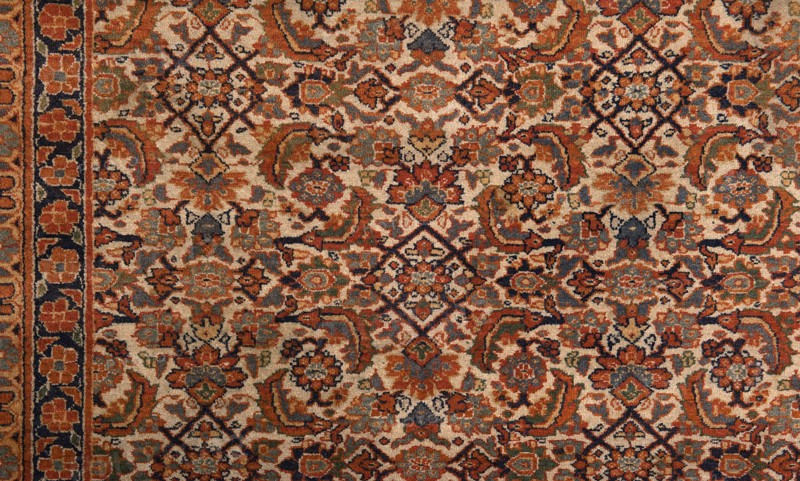 Carpet, Knotted decorative fabric carpet, 19th century, Iran. In the central field on a light cream background: a fine net ornament Herat sack composed of lanceolate leaves, rosettes, palmettes, stylized flowers, and rhombuses. Five-band border with floral motifs; the widest central band on a navy blue background. Colors: brick red, red, green, blue, navy blue, and white. Height 520 cm, width 395 cm., A patterned textile with an ornate repeating design dominated by shades of rust, beige, navy, and muted green, featuring floral and geometric motifs. The surface shows a dense arrangement of stylized flowers, curved leaf-like shapes, and diamond-shaped elements, with darker navy outlines accenting the patterns. Along the left edge there is a vertical border strip with a row of rounded floral shapes on a darker background. The overall texture appears woven or tufted, with subtle variations in color saturation across the design.