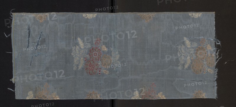 Fabric fragment, 17th century, Italy. Brocaded and morayon grosgrain silk. Weft grosgrain construction with blue silk warp and weft; pattern brocaded with silk threads held with a weft weave in dark pink, beige, salmon, white, and a gold-plated thread braided on a white silk core moriated. Design: loosely scattered smaller and larger floral sprigs on a blue background; stems and leaves in white with petals outlined in red and salmon-pink and filled with gold or left plain; probably larger bushes in two alternating-row types with smaller elements between. Motif size reported 14 x 10.5 cm; preserved fragment dimensions height 57 cm, width 24 cm; selvedge 0.4 cm wide blue and beige silk threads. Weaving edge: 0.4 cm wide silk with blue and beige stripes., A rectangular piece of worn woven fabric lies flat against a dark background, showing a muted blue-gray ground with visible vertical and horizontal weave texture and areas of fraying along the edges; several floral motifs in faded gold, ...