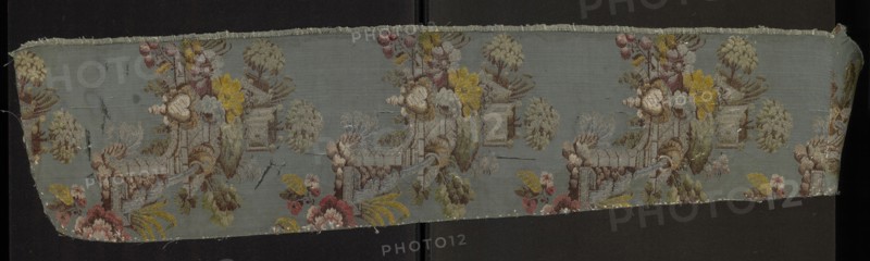 Fabric fragment, silk fabric, patterned, preserved selvedge, Taffeta background in blue-green, brocaded with colorful threads tied in a twill weave. Brocade colors: light and dark greens, browns, pale pinks, dark cherry, blue, gray, silver, olive green, lemon green, reddish brown. Floral motifs and fragments of garden architecture. Dimensions: length 26 x ?, edge width 0.5 cm, height 111 cm, width 21 cm. 1730s, France., A narrow strip of woven fabric with a muted blue-gray background displaying a repeating decorative scene in soft, faded colors; the design includes a group of stylized floral and leafy forms in shades of yellow, pink, red, green, cream, and brown, along with architectural elements resembling a low wall or terrace worked in pale gray and brown tones, and rounded tree-like shapes in pale green and beige. The fabric shows frayed edges and small loose threads along the top and bottom, with areas of slight discoloration and faint surface marks in darker gray and brown. The p...