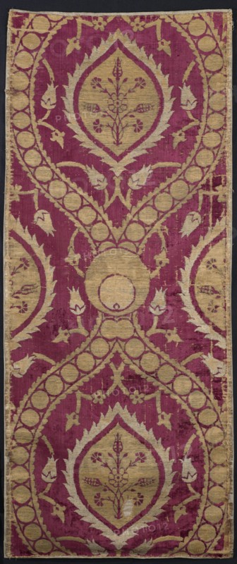 Fabric piece, Repossession, Turkish fabric, 16th century. Red sheared velvet ground with cream and silver patterns in satin and weft weaves. Large repeat of pointed ovals formed by wide ribbons fastened with a peacock's eyes motif; ribbons filled with single circles. Inside ovals: palmettes with a central bouquet of flowers. Surrounding the ovals: winding stems of small flowers. Height 154 cm, width 63 cm., The image shows a vertical textile with a repeating ornamental pattern in deep magenta and muted gold tones on a dark background border. The design features a series of large teardrop-shaped medallions with pointed leafy outlines containing floral motifs, connected by curved bands decorated with a row of circular elements. Stylized leaf and bud shapes extend from the bands and fill the surrounding field. The textile surface shows areas of wear and slight discoloration where the magenta and gold threads vary in intensity, and the edges appear slightly frayed.