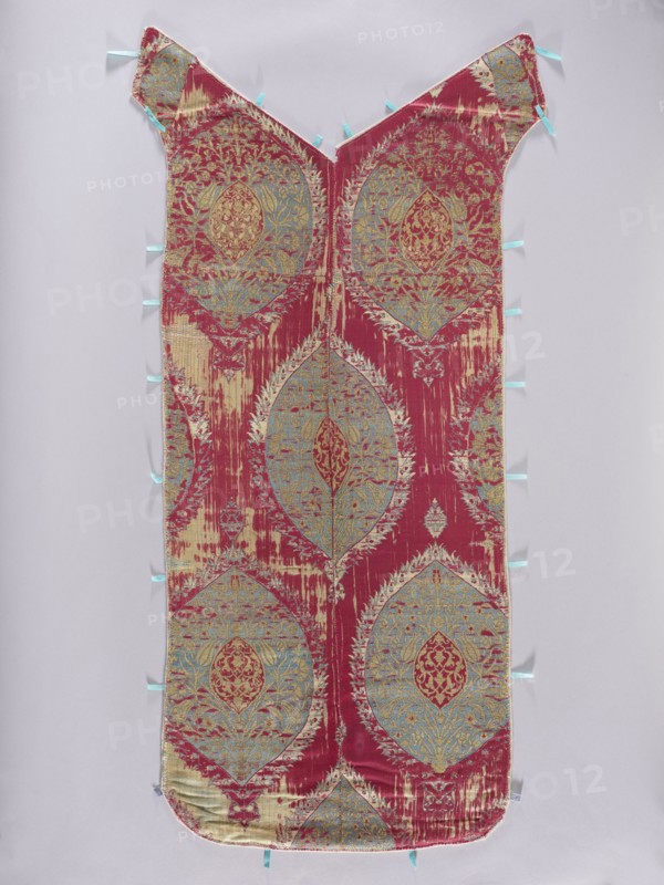 Back of the chasuble, fabrics, metal threads, liturgical vestments, Turkish fabrics, fragments of silk fabrics, patterned fabrics, flowers, leaves, plant ornaments, church paraments, sacred art, churches and chapels furnishings. The back of the chasuble is sewn from two fragments of fabric with motifs of large palmettes placed on a plain red background. The palmettes with serrated edges are decorated inside with floral sprigs and fruit spreading symmetrically and enclosing a smaller palmette with arabesque patterns in the center. Patterns in cream, blue, and gold. Three pieces of fabric sewn into one. Regular plant ornament, unfolding vertically on a red background. Colors: gold, white and sapphire. 17th century, Turkey, liturgical vestment, length 118 cm, width 49 cm back., A long, narrow textile panel with a V-shaped top and rounded bottom, displayed flat against a pale background, showing a repeating ornate medallion pattern in a dominant deep red with accents of light blue and pale...
