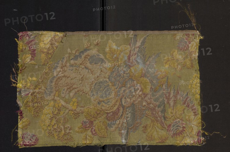 Fabric fragment, fabrics, fragments of silk fabrics, patterned fabrics, three-dimensional patterns, plant patterns. Background: green grodetur cannetillé; brocade pattern: yellow, olive, light and dark pink, green, salmon, white, brown, metal thread with silver braiding. In the Louis XVI style, fruit and flowers on a green background, 18th century, France; fabrics; height 44 cm, width 27 cm., A rectangular textile swatch with a woven floral and foliate design sits against a dark background; the fabric shows a palette of olive green, mustard yellow, muted gold, dusty blue, pale beige, soft pink, and touches of faded red, with areas of worn fraying at the edges and corners, a folded triangular patch at one corner, and a central cluster of stylized flowers and leaves rendered in layered threads that create textured, raised motifs across the surface.