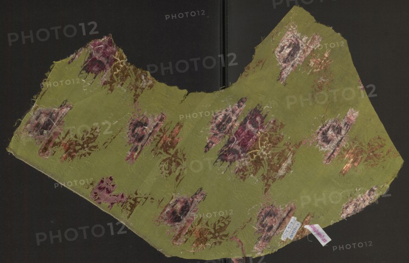 Fabric fragment, silk, unknown origin, 18th-century silk fragment. Pattern: symmetrical composition of flower bouquets and a lace ribbon. Ground: grass-green background grosgrain weave with a delicate, fanciful drawing of clouds—leaves achieved by releasing weft threads. Embroidery/broaching colors: blue, olive, white, pale pinks and reds, cherry red, cream, salmon, black. Measurements: reported 29.5 x ?; selvedge width 0.6 cm; overall height 32 cm, width 77 cm. Date: second half of the 18th century; possible Russia., A worn piece of fabric with an irregular curved shape lies against a dark background, predominantly olive green with patches of faded pink, burgundy, brown, beige, and cream forming floral and abstract motifs; the fabric shows frayed edges and loose threads, areas of abrasion and thinness, and several clusters of brushy, textured color where the weave is disturbed, while a small folded cloth tag of pale color is tucked near one edge.