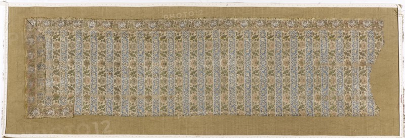 Fragment of a belt with a continuous pattern of alternating silhouetted tendrils with an iris motif and colorful tendrils with saxophone leaves and symmetrical palmettes. Border of floral tendrils and medallions with portraits of men in turbans. Background of the continuous belts: navy blue and gold-lipped. Background of the border belt: gold-lipped; background in the cartouche with a male bust – silver-white. The silhouetted motifs in the shelves are similar to patterns in other contemporary belts. The warp is made of salmon-colored silk yarn; the weft is gold metal thread wrapped around a silk thread. Preserved only in fragments; part of the hem was cut from the body and sewn along the length of what was likely a severed head. First half of the 17th century, Iran, clothing accessory; height 115 cm, width 32 cm., A rectangular textile fragment with a decorative striped pattern laid on a plain tan backing; the fragment shows alternating vertical bands of floral motifs in muted blue, gr...