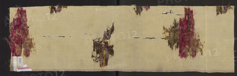 Fragment of fabric, patterned silk fabric with preserved selvedge, floral patterns, 18th-century French silk. Description: white cream background - gros de Tours liseré, brocaded with threads in olive green, dark green, dark pink, pale red, heather. Inscription/label: Gros de tours liseré, 1 lat de lancé, broché a liagé repris. Dimensions: height 81 cm, width 23 cm; fragment 59 x ?; selvedge width 0.6 cm., A long rectangular piece of pale beige fabric with a textured weave lies against a dark background; there are several torn and frayed floral patches in shades of deep red, burgundy, brown, olive green, and muted yellow scattered across the surface, including a tall vertical distressed patch near one short edge, a similar vertical patch near the opposite short edge, and a smaller vertical cluster near the center, with a few smaller fragmented floral fragments near the top edge; thin horizontal dark navy lines and small dark smudges interrupt the beige ground in places, and loose fiber...